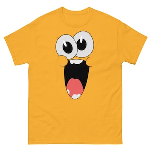 Könnte beinhalten: Ein gelbes T-Shirt mit einem Comic-Gesicht auf der Vorderseite. Das Gesicht hat große Augen, einen weit geöffneten Mund und eine rosa Zunge.