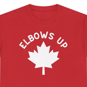 Puede incluir: Camiseta roja de manga corta con una hoja de arce blanca y el texto "ELBOWS UP" impreso en la parte delantera.
