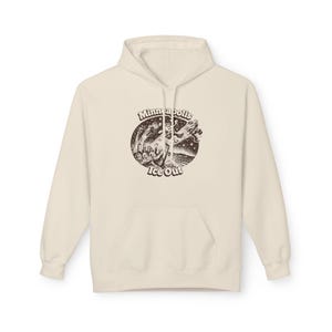 Puede incluir: Sudadera con capucha color crema con bolsillo delantero y capucha con cordón. La parte delantera presenta un gráfico circular marrón con las palabras "Minneapolis Ice Out" y una escena invernal.