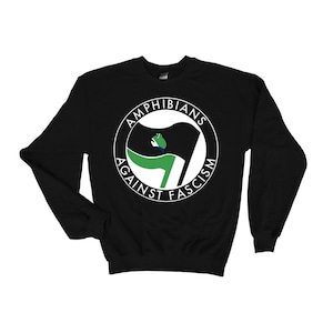 Puede incluir: Sudadera negra con un diseño circular que dice "AMPHIBIANS AGAINST FASCISM". El diseño presenta una bandera estilizada con elementos verdes, negros y blancos, y una silueta de rana. La sudadera es de cuello redondo.