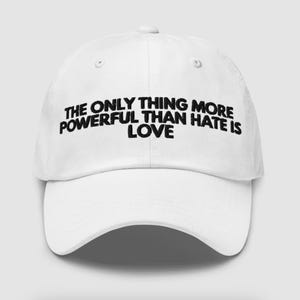 May include: White baseball cap with a curved brim. The cap features the embroidered text: "THE ONLY THING MORE POWERFUL THAN HATE IS LOVE" in black, centered on the front panel. The cap has a classic six-panel design with a button on top.
