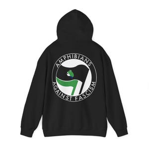 Puede incluir: Sudadera con capucha negra con un diseño circular blanco en la espalda. El diseño incluye el texto "AMPHIBIANS AGAINST FASCISM" y un motivo de bandera con elementos verdes y negros.
