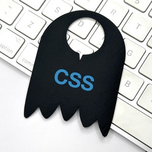 Könnte beinhalten: Ein schwarzer, geisterförmiger Türhänger mit dem Wort "CSS" in blauen Buchstaben auf einer weißen Tastatur.