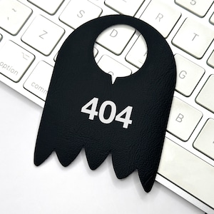 May include: A black, ghost-shaped door hanger with a hole at the top and the number 404 printed in white on the front. The hanger is lying on a white keyboard.