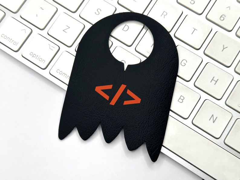 May include: Black leather door hanger shaped like a ghost with a red and white code symbol, "</>", on the front. The hanger is hanging on a white keyboard.