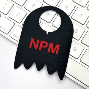 Puede incluir: Una etiqueta de cuero negro con un agujero recortado en la parte superior y la palabra "NPM" en letras rojas. La etiqueta está sobre un teclado blanco.