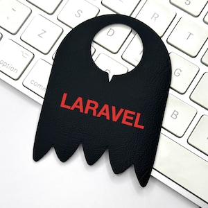 May include: A black, bat-shaped tag with the word "LARAVEL" in red lettering, resting on a white computer keyboard. The tag has a hole at the top and a jagged bottom edge. The keyboard keys are white.