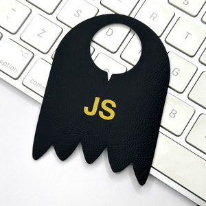 May include: Black leather door hanger with a jagged bottom edge and a cut-out circle at the top. The hanger has the initials "JS" in yellow lettering.