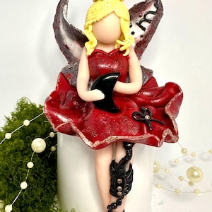 May include: A fairy figurine in a red dress with blonde hair and black details. The fairy holds a black crescent moon and has black embellishments on her legs. The wings are grey and red.