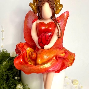May include: A handmade figurine of a woman in a red dress and orange skirt, holding a red object. The figurine has golden ram horns and wings. The figure is on a white base, with green moss and pearl accents.