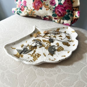 Peut inclure: Plat en céramique blanche en forme de feuille avec des accents dorés. Le plat est rempli de bijoux, y compris des boucles d'oreilles, une bague et un collier.
