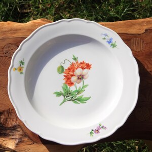 Könnte beinhalten: Weißer Keramikteller mit gewelltem Rand, mit einem Blumendesign mit orangefarbenen, weißen und grünen Elementen. Kleine florale Akzente in Gelb, Blau und Lila sind um den Rand verstreut. Der Teller liegt auf einer Holzoberfläche.