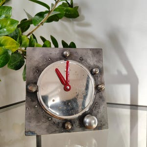 May include: A square, industrial-style clock with a silver face and red hands. The clock is set in a metal frame with various silver and bronze embellishments. The clock's hands point to approximately 2:00. The clock is displayed on a glass surface.