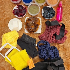 May include: A collection of natural dye materials and accessories. The image shows powders in red, blue, green, and orange, plant-based dye ingredients, and various scrubbers and wraps. A pink spray bottle is also visible.