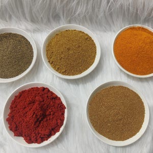 Pode incluir: Cinco tigelas brancas cheias de especiarias diferentes estão dispostas sobre uma superfície branca texturizada. As especiarias incluem pimenta preta, caril em pó, paprika, açafrão e uma especiaria castanha. As especiarias são finamente moídas e têm texturas e cores variadas.