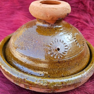 Puede incluir: Una olla de tagine de cerámica esmaltada con tapa abovedada y base circular. La olla es principalmente marrón con patrones florales decorativos en la tapa. La tapa tiene un pequeño asa de terracota sin esmaltar. El tagine está sobre una superficie roja.