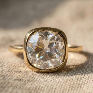 May include: A gold ring featuring a large, clear, cushion-cut diamond. The diamond is set in a bezel setting, and the band is a simple, polished gold. The ring is displayed on a neutral-colored, textured surface.