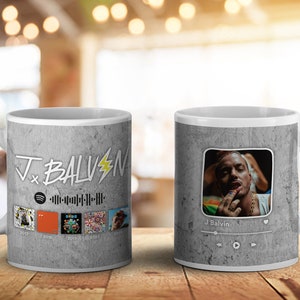 May include: White ceramic mug featuring a gray textured design with the text "J x BALVIN" and a Spotify logo. The mug displays album art and a photo of J Balvin with a music player interface.