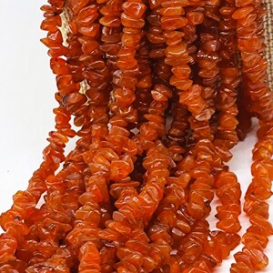 Op de afbeelding: Close-up van strengen oranjerode edelsteen kralen. De kralen zijn onregelmatig van vorm en aan elkaar geregen, waardoor een watervaleffect ontstaat. De edelstenen hebben een warme, doorschijnende kleur en lijken carneool te zijn.