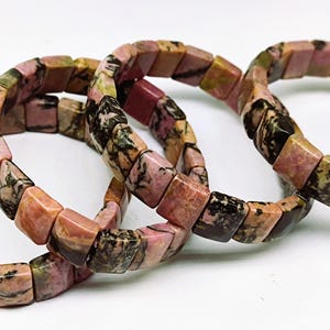 Op de afbeelding: Vier edelsteenarmbanden. Elke armband is gemaakt van vierkant geslepen, gepolijste stenen in tinten roze, bruin en groen. De armbanden zijn in een cirkelvorm gerangschikt, waarbij de natuurlijke variaties in de steen worden getoond.