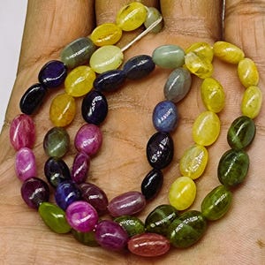 May include: A strand of oval-shaped gemstone beads in various colors, including yellow, green, blue, pink, and purple. The beads are strung on a clear thread and are arranged in a random pattern.