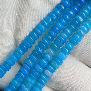 May include: A strand of light blue opal beads, with a slight iridescent shimmer, arranged in rows on a white fabric background.
