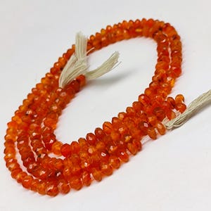 May include: A collection of orange carnelian beads, each faceted to catch the light. The beads are strung on a light-coloured string, suitable for a necklace or bracelet. The polished surface of the beads gives a warm, vibrant appearance.
