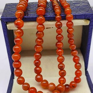 May include: A strand of carnelian beads in a blue velvet box. The beads are a deep orange color and are round in shape.