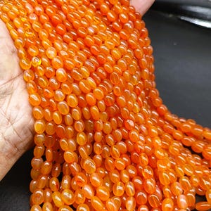 May include: A collection of vibrant orange carnelian gemstone beads. The oval-shaped beads are strung together, creating a flowing, textured appearance. The gemstones have a polished, glossy finish, reflecting light and showcasing their natural colour variations.