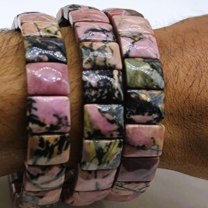 Op de afbeelding: Drie armbanden gemaakt van rechthoekige, gepolijste stenen. De stenen zijn voornamelijk roze met zwarte, groene en grijze adering. De armbanden worden om een pols gedragen, waarbij de natuurlijke kleurvariaties en patronen van de stenen worden getoond.