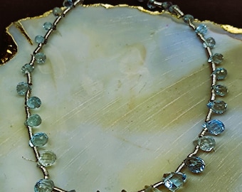 May include: A strand of light blue teardrop-shaped gemstone beads strung on a silver wire. The beads are arranged in a circle on a white and gold surface.