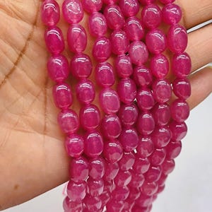 May include: Strands of polished, oval-shaped, fuchsia-coloured beads. The beads are strung together, and held in a hand. The beads have a glossy finish and are uniform in size and shape. Ideal for jewelry making.