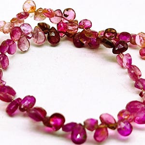 May include: A strand of pink and orange gemstone beads. The beads are faceted and have a teardrop shape. The beads are strung on a clear thread.