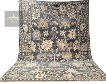 Puede incluir: Una alfombra grande de color gris oscuro con un patrón floral en beige y crema. La alfombra tiene un borde con un diseño geométrico.