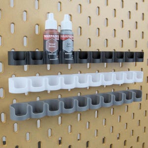 Pode incluir: Um painel perfurado com três fileiras de suportes para frascos de tinta em preto, branco e cinza. Dois frascos de tinta estão na fileira superior. Os frascos estão rotulados como "Dryad Brown" e "Ash Grey". O painel perfurado tem orifícios ovais espaçados.