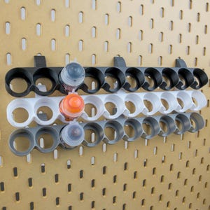 Peut inclure: Support à bouteilles de peinture à trois niveaux en noir, blanc et gris, monté sur un panneau perforé. Chaque niveau contient plusieurs bouteilles, avec quelques bouteilles visibles dans les supports. Le panneau perforé est de couleur marron clair.