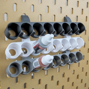 May include: Three-tiered paint bottle holder in gray and white, mounted on a pegboard. Each tier holds multiple bottles, with two bottles visible. The holders are designed to keep paint bottles organized and accessible.