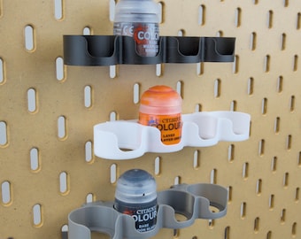May include: Three-tiered paint bottle storage system in black, white, and gray. Each tier holds Citadel Colour paint bottles. The storage system is mounted on a pegboard with evenly spaced holes. The paint bottles are various colors.