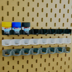 Può includere: Una pannello forato con tre piccoli flaconi di vernice gialla, blu e nera, tenuti da supporti neri. Sotto ci sono supporti bianchi e grigi per flaconi di vernice. Le bottiglie sono etichettate "LACQUER PAINTO".