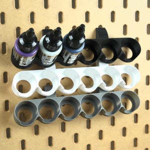 Peut inclure: Support de bouteilles de peinture à trois niveaux en noir, blanc et gris, monté sur un panneau perforé. Le niveau supérieur contient trois flacons de peinture avec le mot "PRO" visible. Les supports sont conçus pour ranger et exposer les flacons de peinture en toute sécurité.