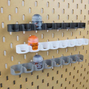 Peut inclure: Trois supports pour flacons de peinture imprimés en 3D, en noir, blanc et gris, montés sur un panneau perforé. Chaque support comporte plusieurs emplacements pour contenir des flacons de peinture. Les supports sont conçus pour organiser et exposer les flacons de peinture.