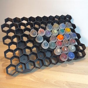 Könnte beinhalten: Ein schwarzer, sechseckiger Lackständer aus Kunststoff mit 25 Lackflaschen in verschiedenen Farben. Der Ständer ist so konzipiert, dass er kleine Lackflaschen aufrecht hält.