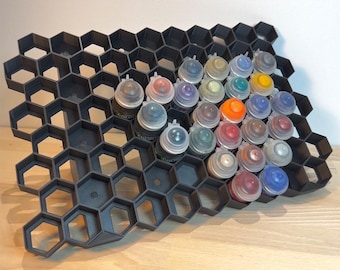 Op de afbeelding: Een zwarte, zeshoekige verfstandaard van plastic met 25 verfpotjes in verschillende kleuren. De standaard is ontworpen om kleine verfpotjes rechtop te houden.