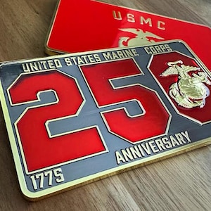 May include: Two rectangular metal plaques. The front plaque is gray with red lettering that reads "UNITED STATES MARINE CORPS 25 ANNIVERSARY 1775 2025". The top plaque is red with "USMC" in gold lettering. Both have gold trim.