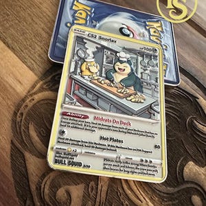 May include: A custom-made trading card featuring a cartoon Snorlax character in a kitchen setting. The card has a yellow border and includes text such as "Midrats On Deck" and "Hot Plates". The card is placed on a wooden surface.