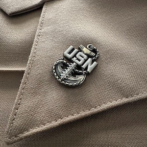 May include: A silver and white US Navy anchor pin with the letters USN on the anchor. The pin is on a brown fabric background.
