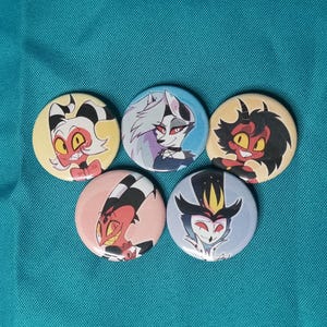 May include: Five round pinback buttons featuring cartoon characters with various designs and colors. The buttons are arranged on a teal fabric background. Each button has a different character design.