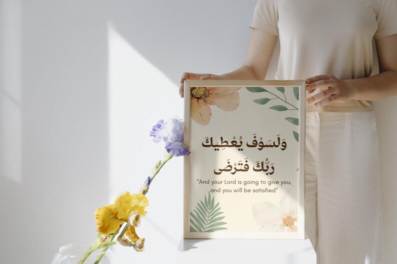 Pu&ograve; includere: Una stampa incorniciata con calligrafia araba e traduzione inglese, con accenti floreali e di foglie in colori tenui. La stampa &egrave; tenuta da una persona. Un vaso di fiori gialli e viola &egrave; in primo piano.