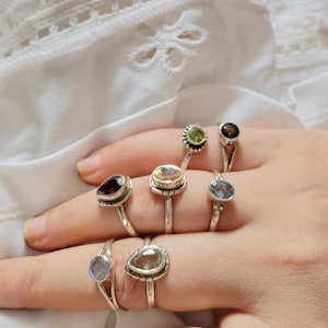 Peut inclure: Une main portant sept bagues en argent avec des pierres précieuses de différentes couleurs. Les bagues présentent des pierres en forme de goutte d'eau dans des tons de vert, bleu, jaune et rouge.