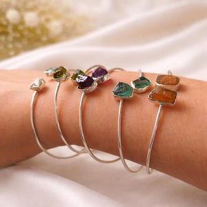 May include: A collection of silver-toned cuff bracelets, each adorned with a unique, irregularly shaped gemstone. The gemstones vary in color, including clear, green, purple, and orange. The bracelets are displayed on a wrist against a soft, white background.